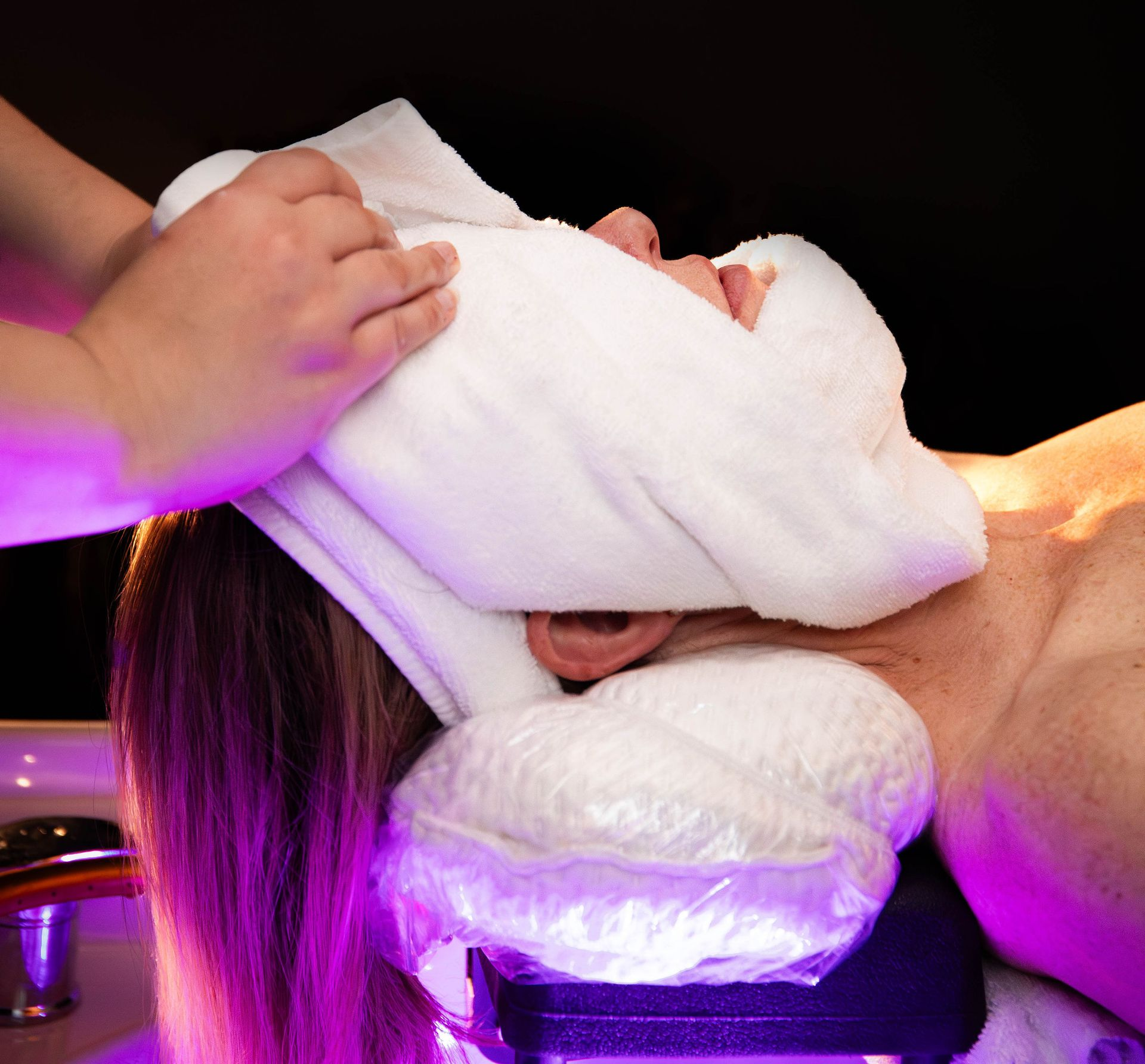 A spa professional holds a warm white towel over a client’s face while they rest on a pillow under purple ambient light.