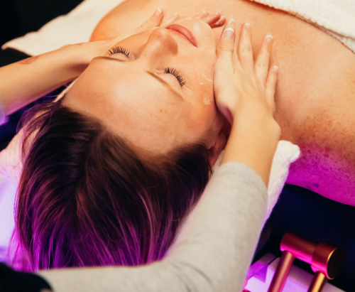 A person receiving a relaxing facial massage in a spa setting with soft, purple-toned lighting.