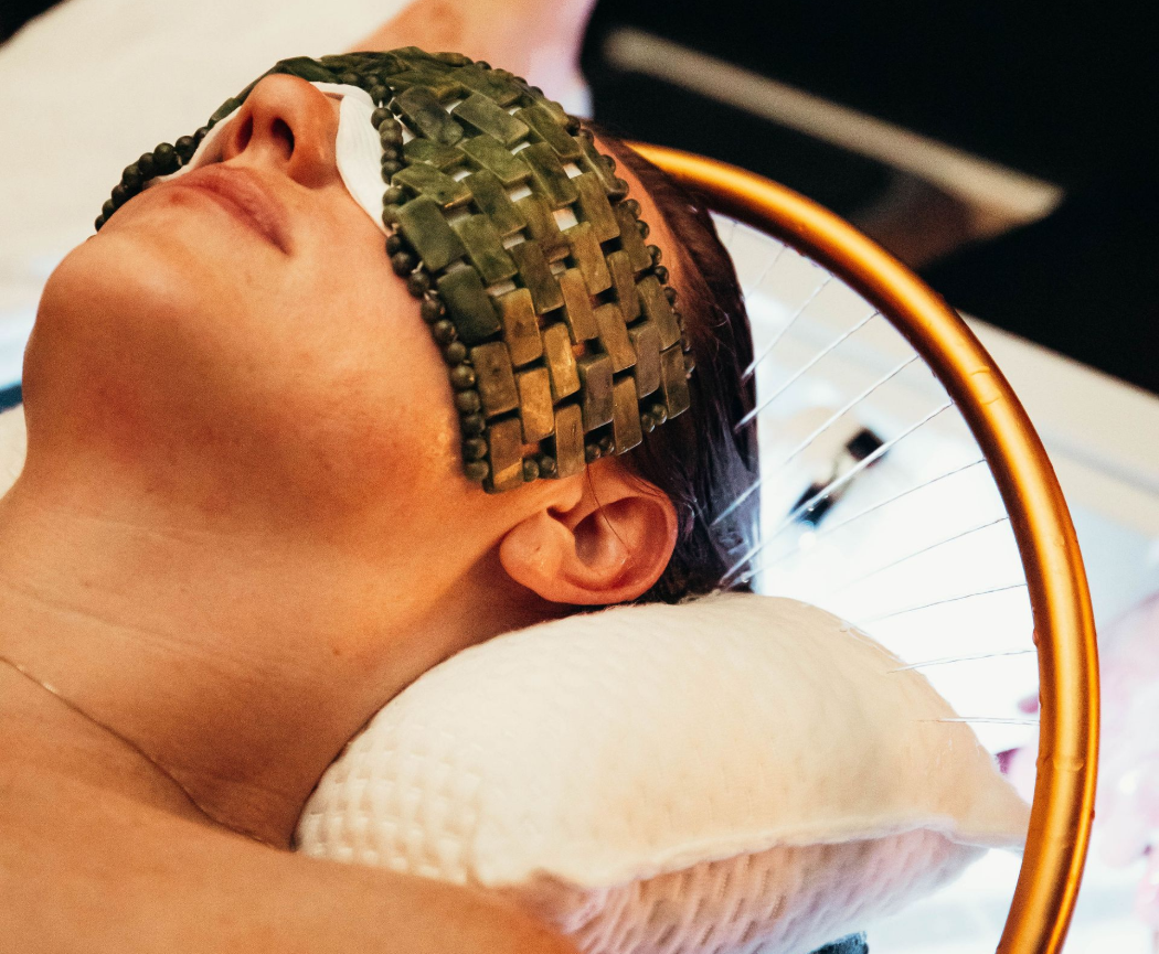 A person lying down with a woven jade stone eye mask resting over their eyes during a spa or facial treatment.