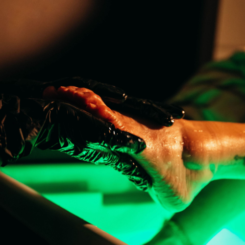 A gloved hand gently touches a bare foot, illuminated by a vivid green light in a dark environment.