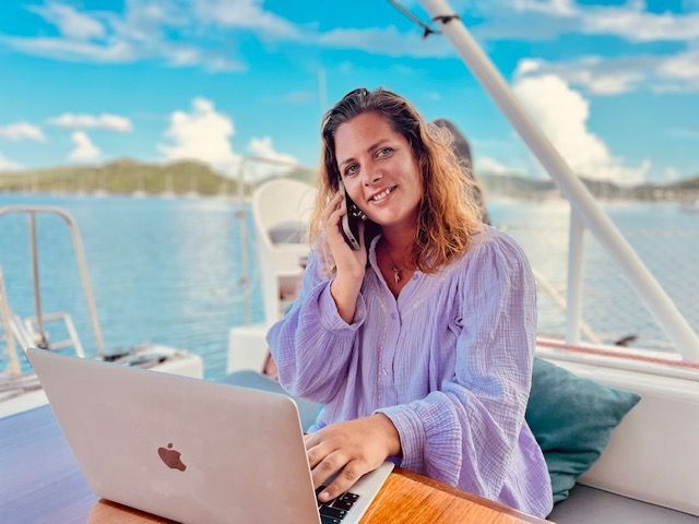 Een vrouw zit op een boot en gebruikt een laptop terwijl ze belt via een mobiele telefoon.