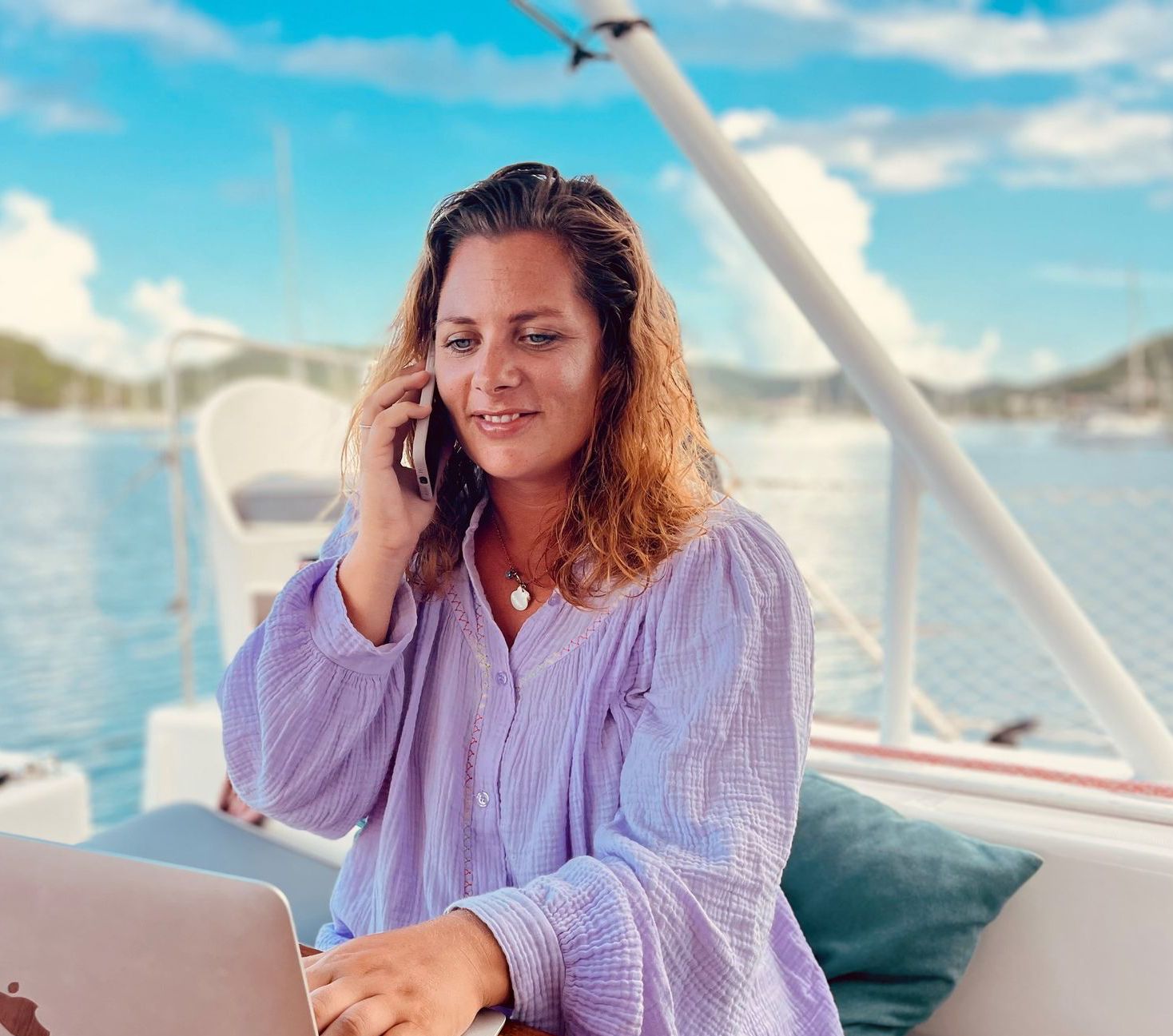 A woman is sitting on a boat using a laptop and talking on a cell phone.