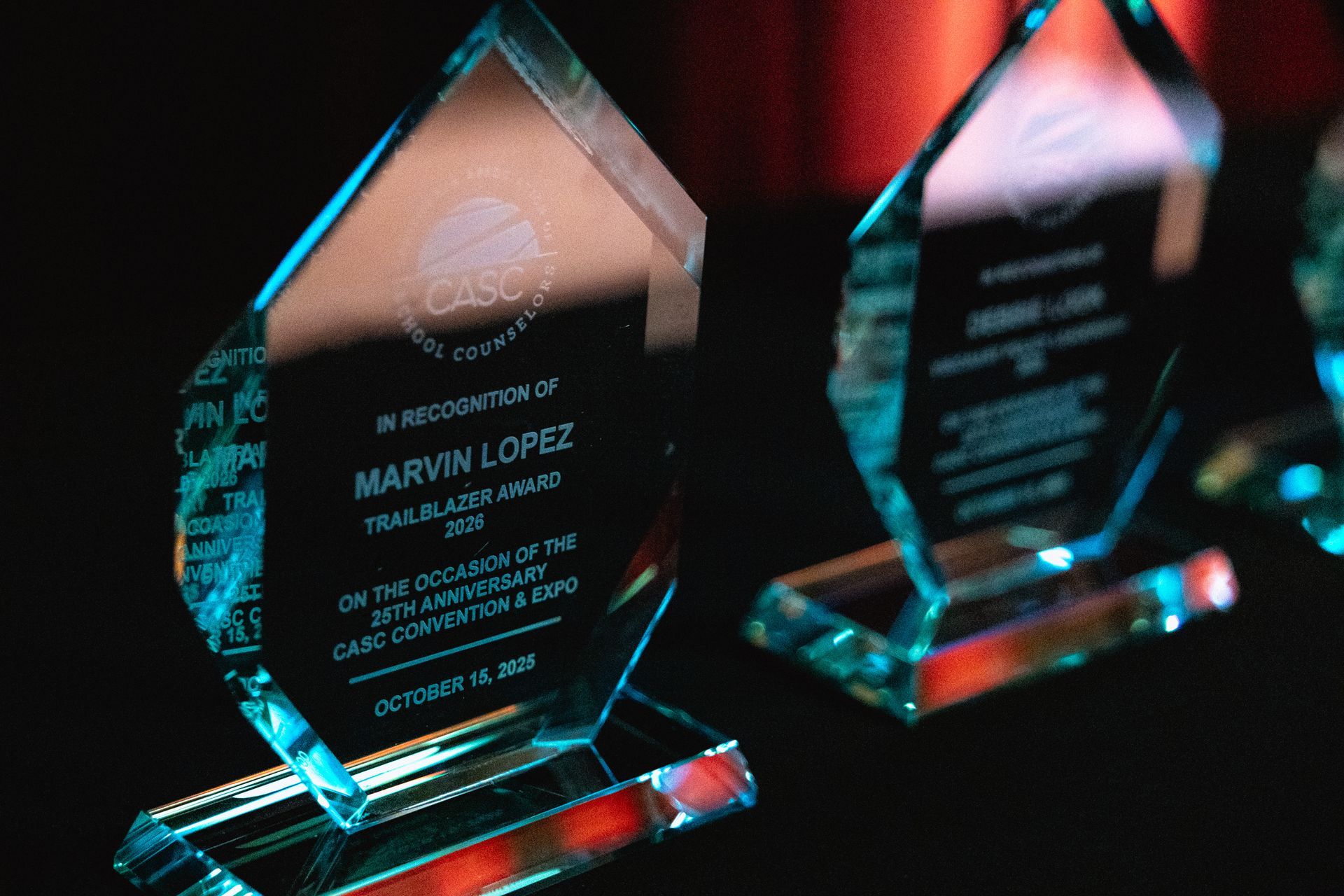 Two glass award trophies on a dark table with red and teal reflections