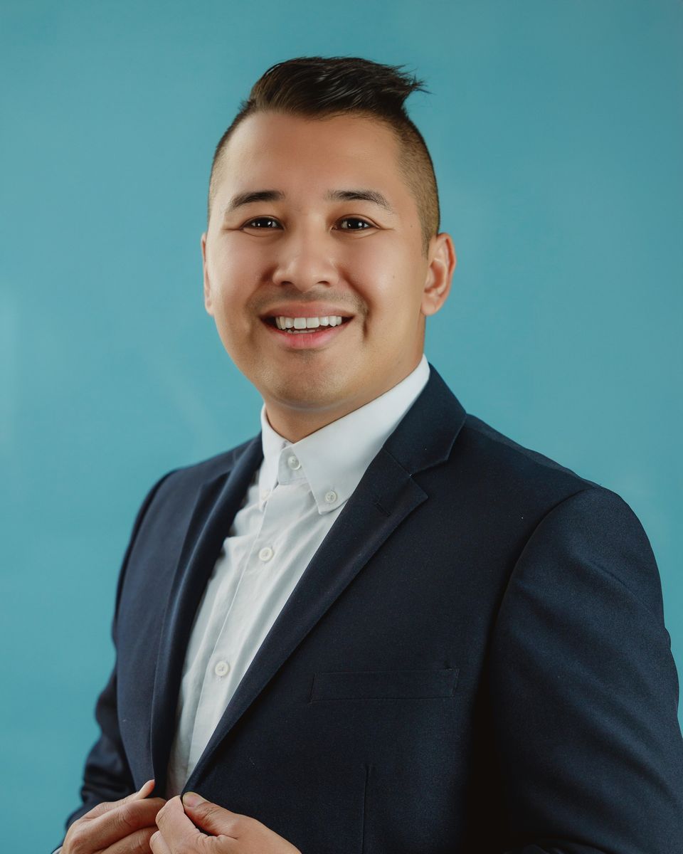 Man in navy suit jacket and white shirt smiles against a teal background.