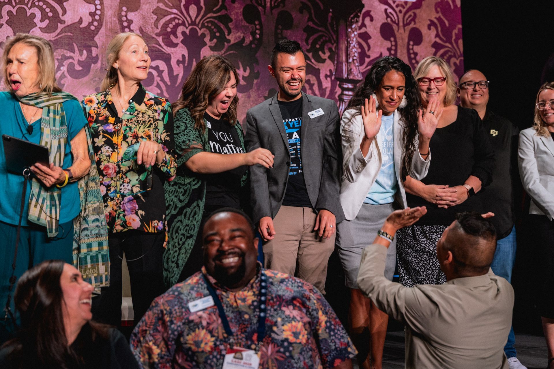 Group of people on stage reacting with surprise and delight. Some are smiling, others have hands up.