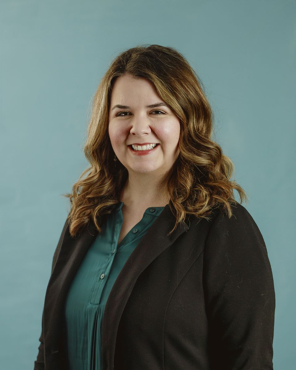 Woman smiling, wearing black blazer and teal shirt, against a blue backdrop.