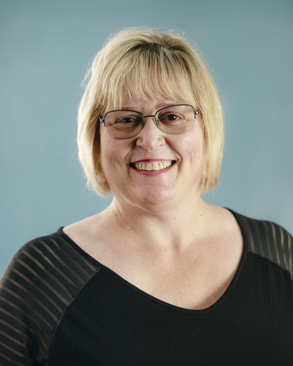Woman with short blonde hair and glasses smiles against a blue backdrop; wearing a black top.