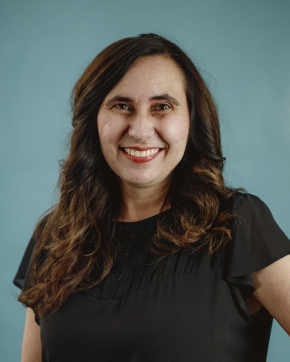 Woman with long brown hair smiles, wearing a black top against a blue background.