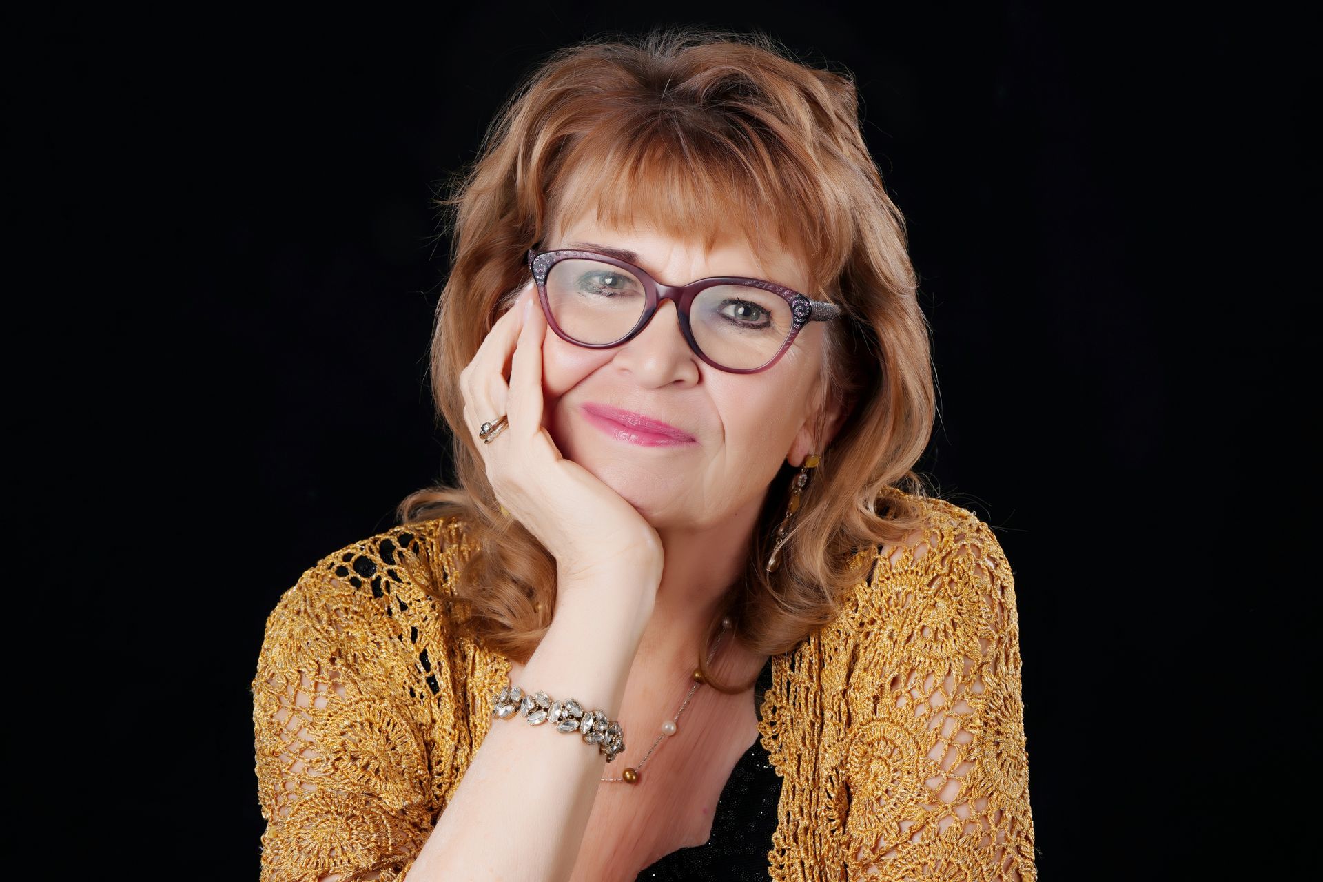 Woman with glasses, resting chin on hand, smiles. Wearing yellow lacy jacket against a black background.