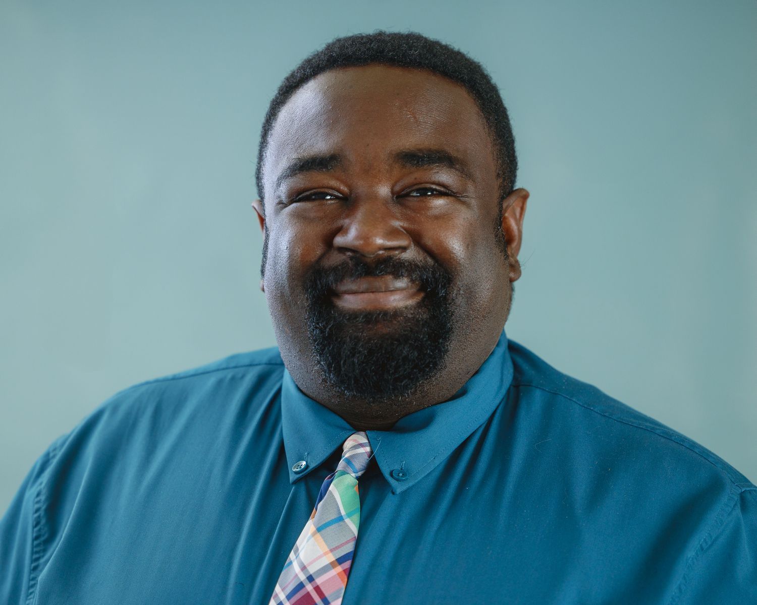 Man with a beard, wearing a blue shirt and patterned tie, smiles in front of a blue background.