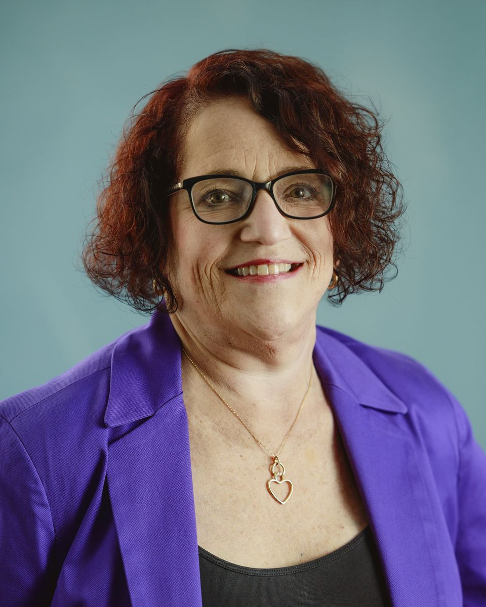 Woman with glasses, short red hair, wearing a purple blazer, black top, and a heart pendant, smiling.