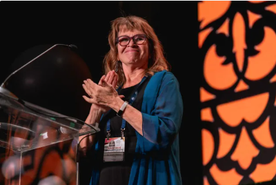 Woman clapping, standing at a podium with an orange patterned backdrop. She is wearing glasses and a blue shawl.