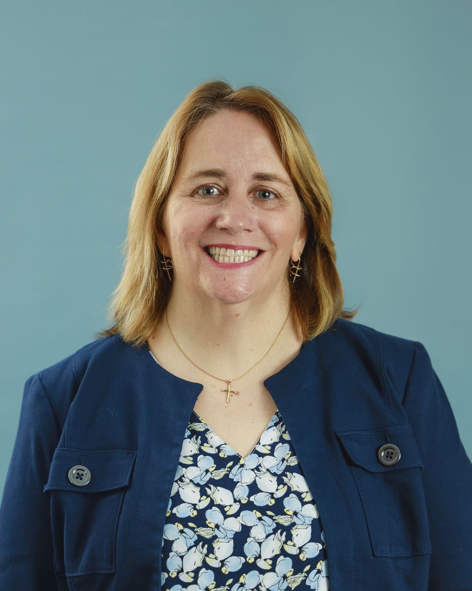 Woman in navy blazer smiles, wearing a patterned top and gold necklace. Against a blue background.