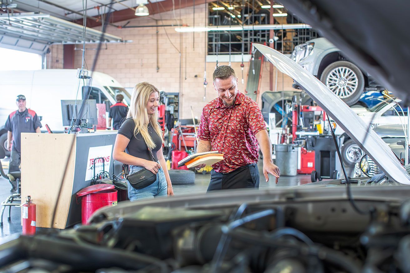A mechanic shows a customer the engine of a car in a garage. Both are smiling, and other workers are visible.
