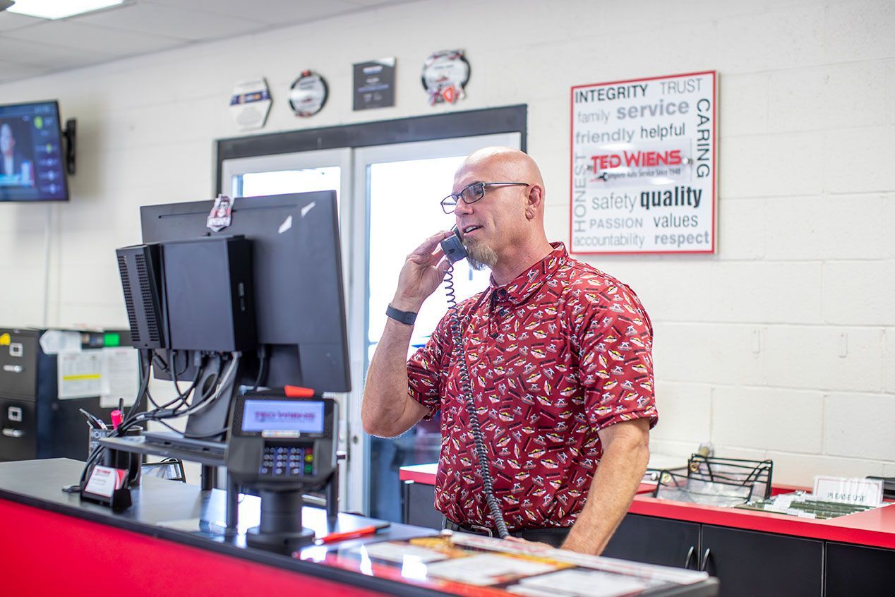 A man in a red floral shirt answers a phone at a service counter. He stands in a shop with a computer monitor and a sign.
