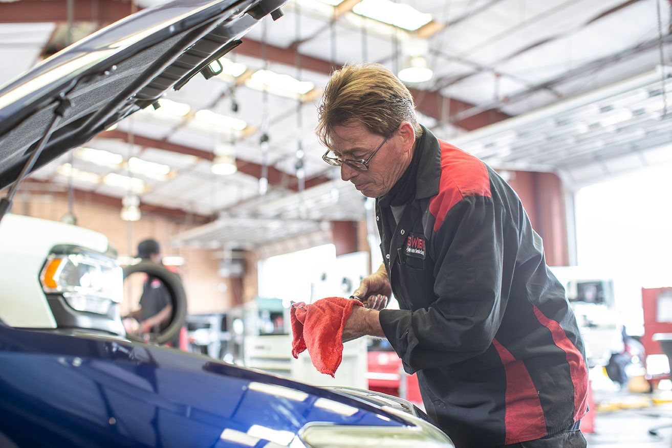 Mechanic in a red and black uniform wiping down a blue car engine in a brightly lit garage.