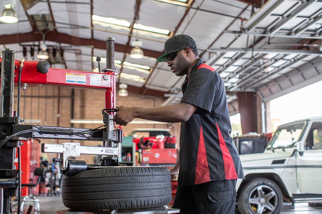 Mechanic in a garage, wearing a hat and uniform, working on a tire with a machine. A luxury SUV is parked nearby.