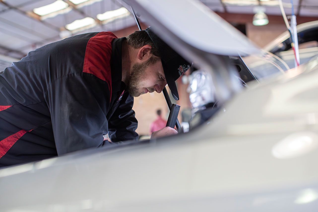 Mechanic in blue and red uniform working on a car engine with the hood up inside a garage.