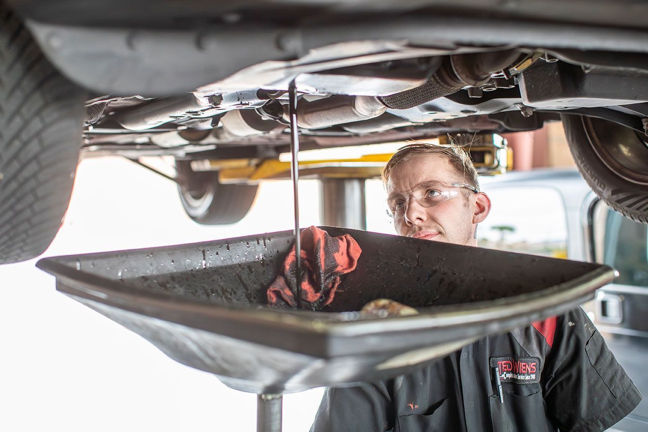 Mechanic draining oil from a car, holding a catch pan. He is wearing safety glasses and is in a garage setting.