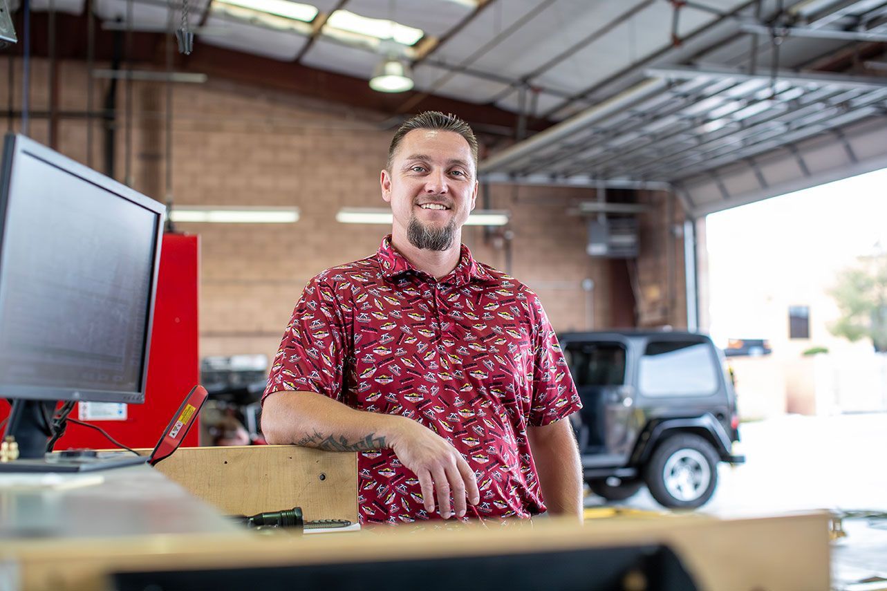 Man in red shirt leans, smiles in a garage. Black Jeep sits behind him.