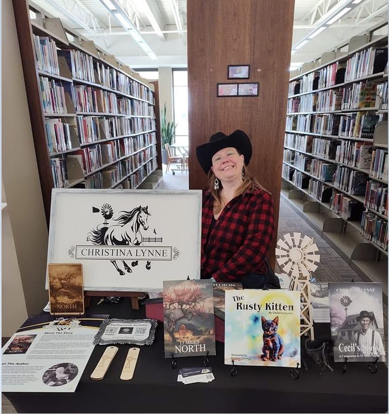 Woman at a book signing, wearing a cowboy hat. Books and a sign with a horse graphic are on the table in front of her.