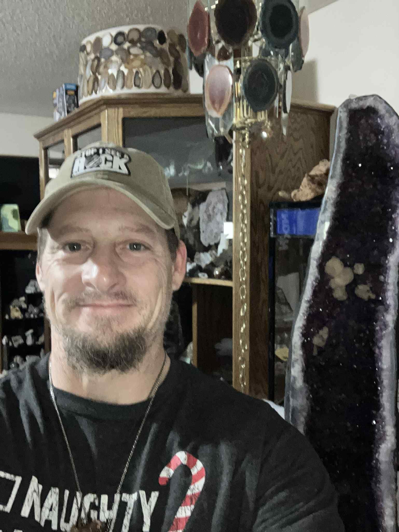 Man wearing a hat and shirt smiling indoors, with a large amethyst crystal and decorative objects.