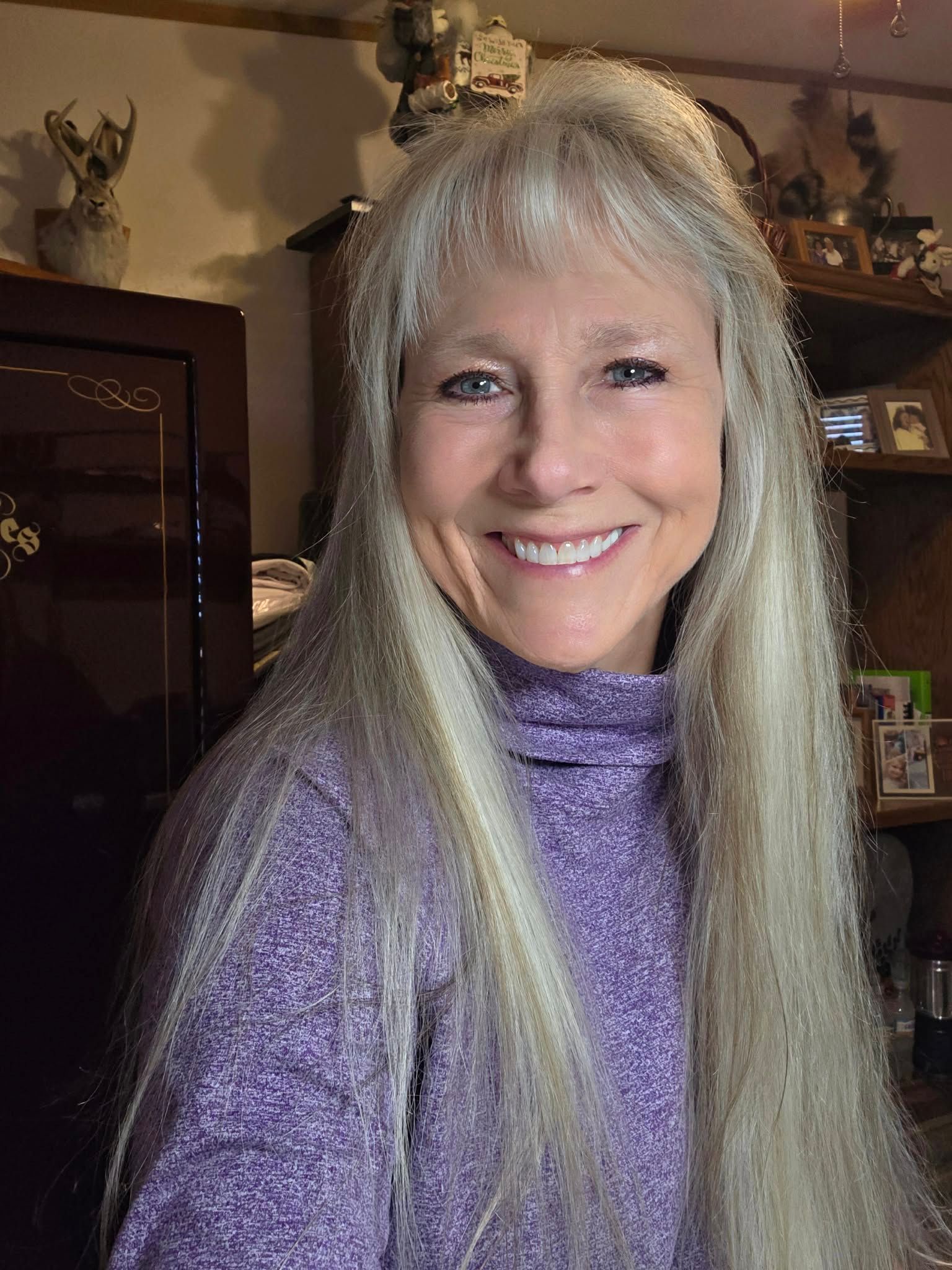 Woman with long gray hair smiles, wearing a purple turtleneck, indoors.