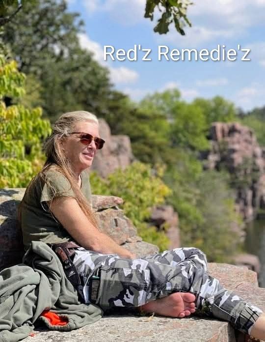 Woman sitting on a rock with bare feet, sunglasses, and camouflage pants, by a body of water. 