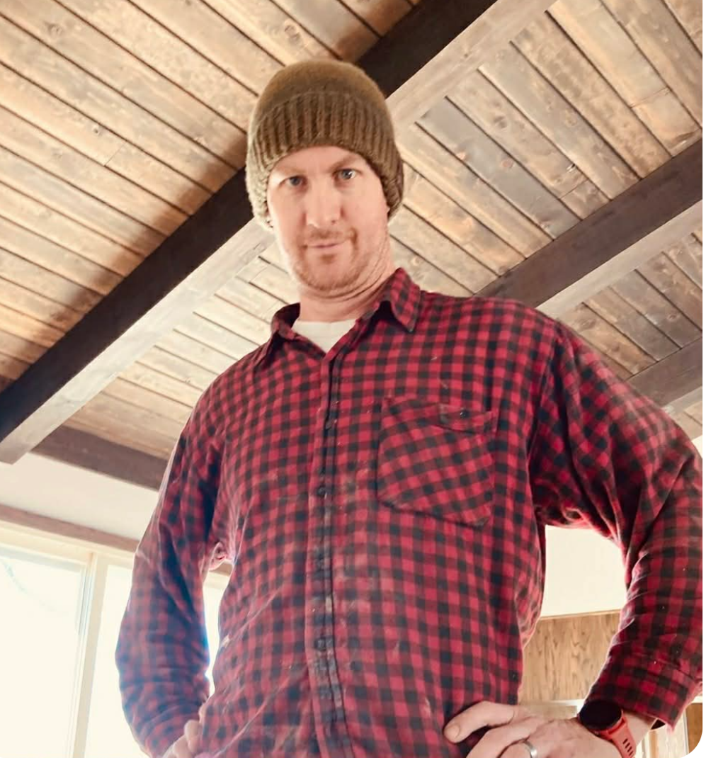 Man in red plaid shirt and brown beanie, arms on hips, looking at the camera. Wooden ceiling in background.