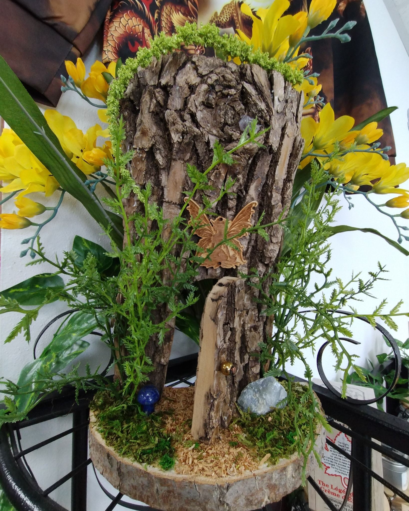 A tree bark sculpture with greenery, yellow flowers, moss, and small rocks on a shelf.
