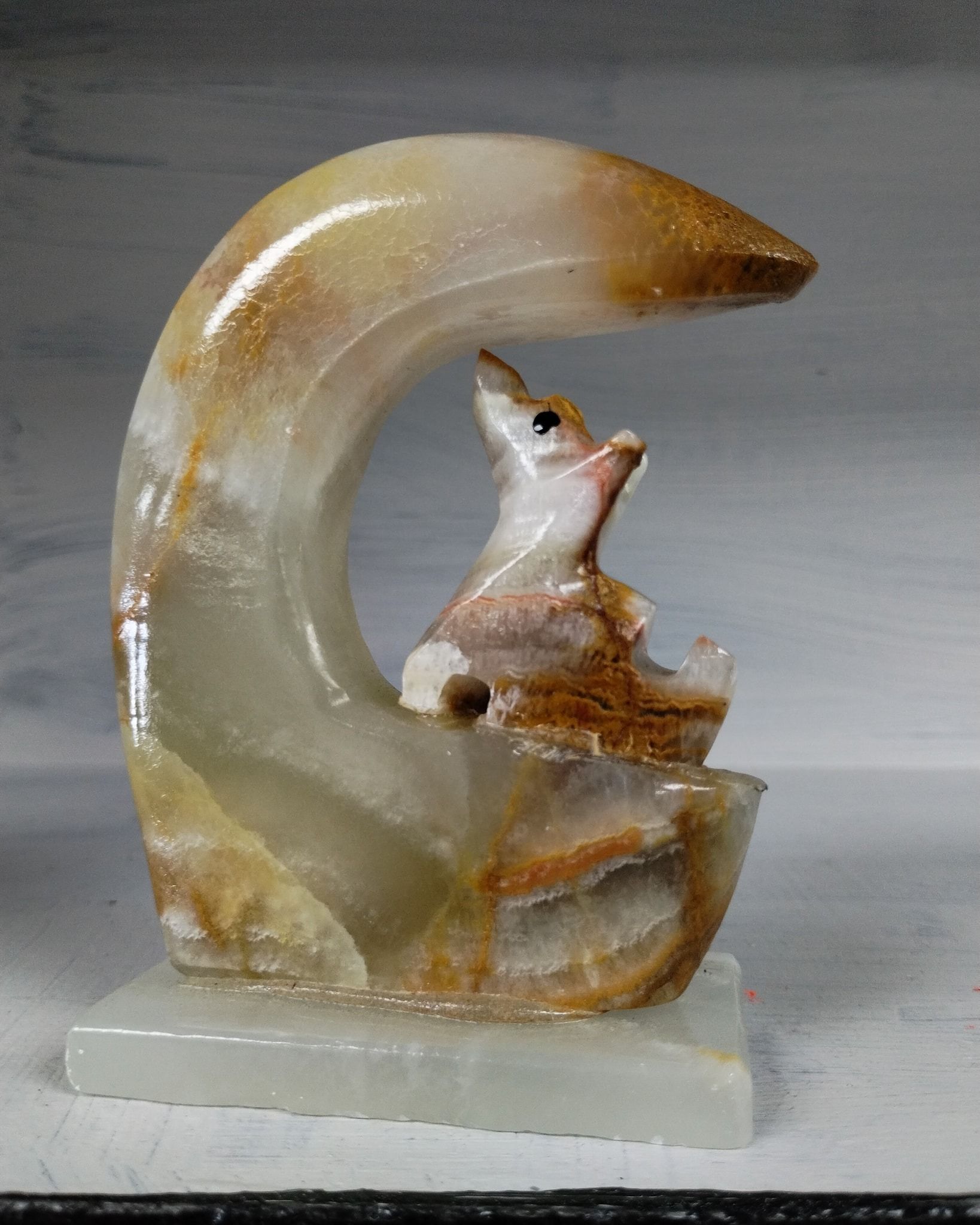 Sculpture of a fox-like creature looking up, sheltered by a curved, moon-shaped onyx. Set on a white base.