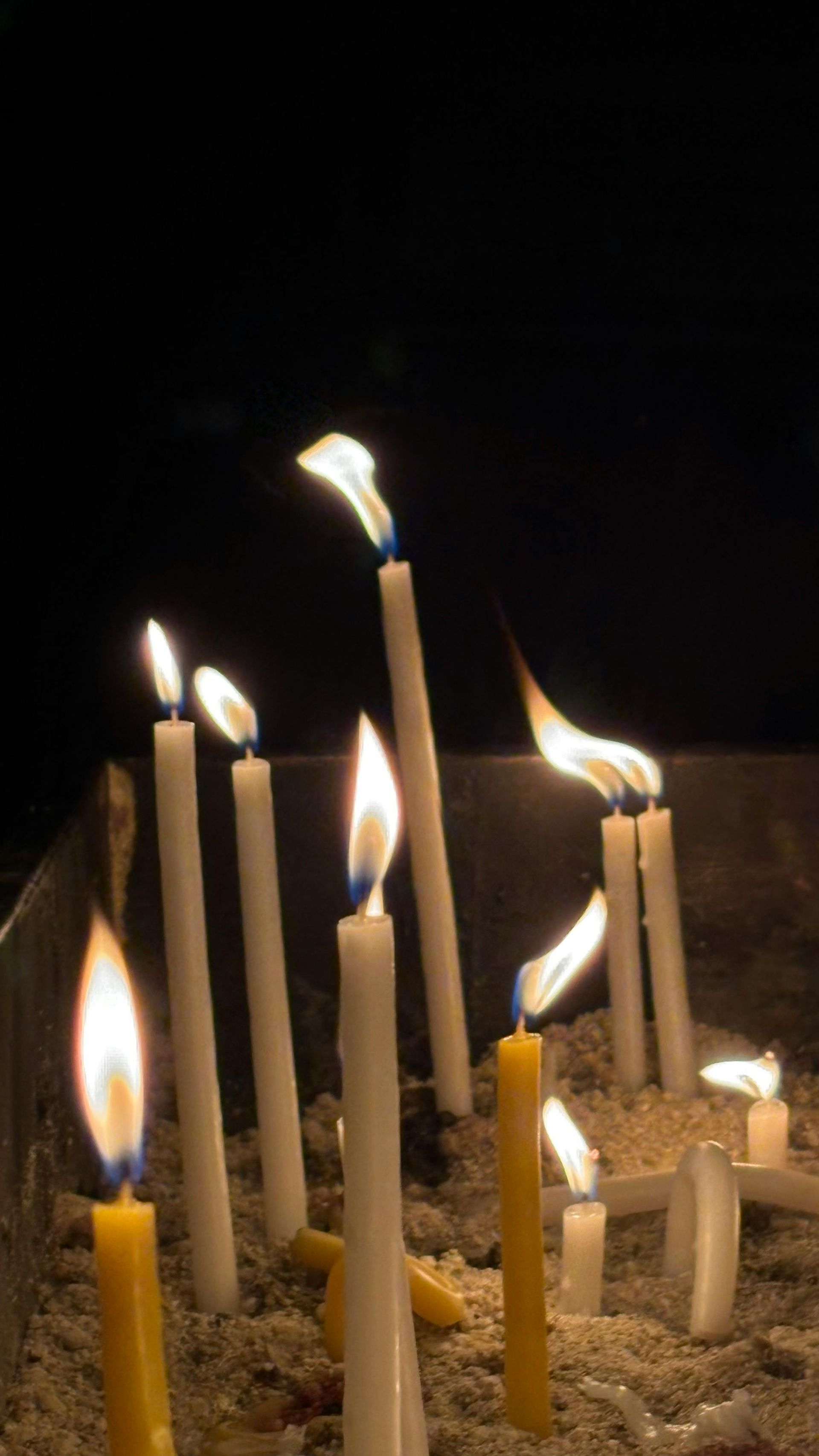 Burning white and yellow candles in sand, flames flickering against a dark background.