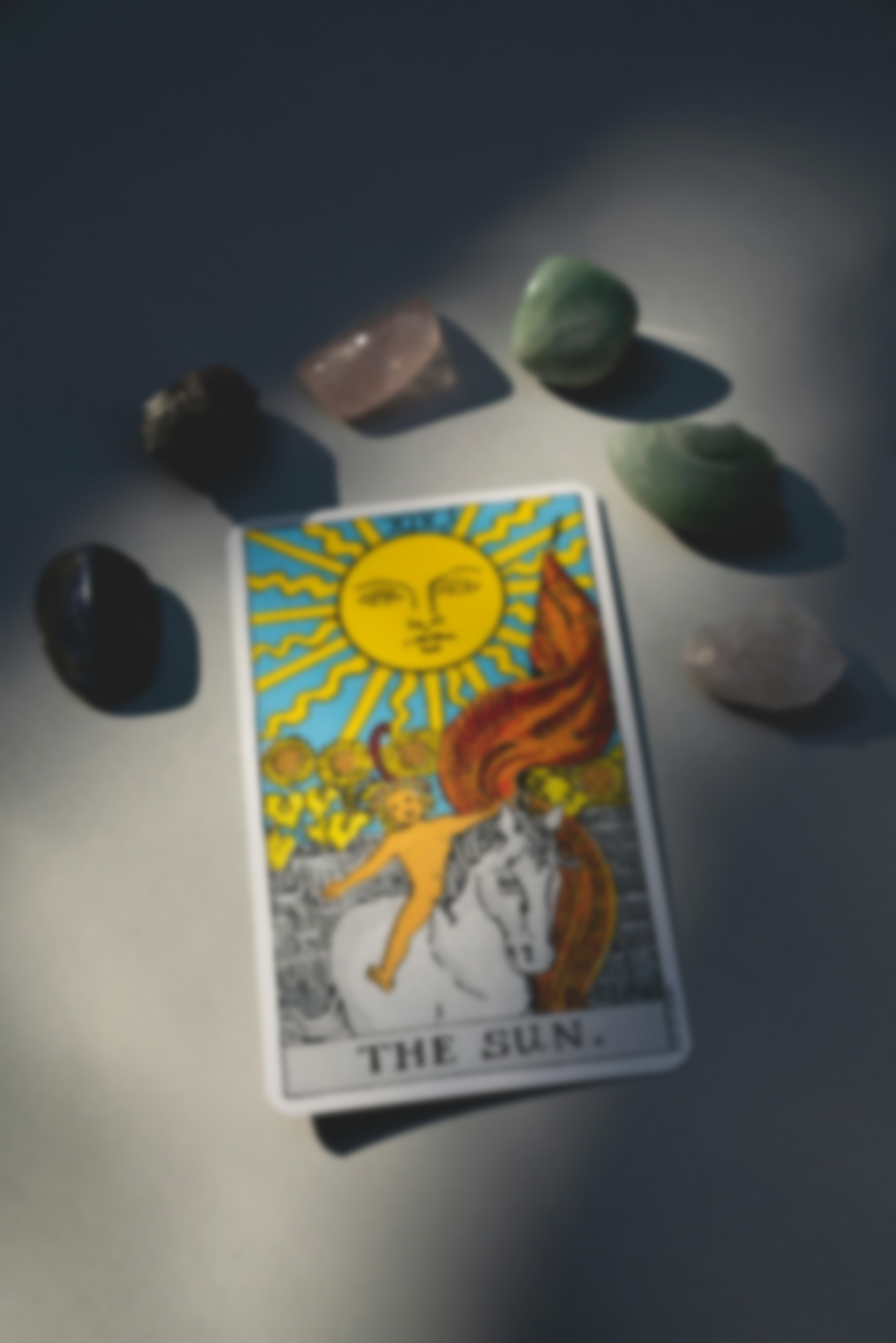 The Sun tarot card surrounded by crystals on a neutral surface. The card depicts a child on a white horse.