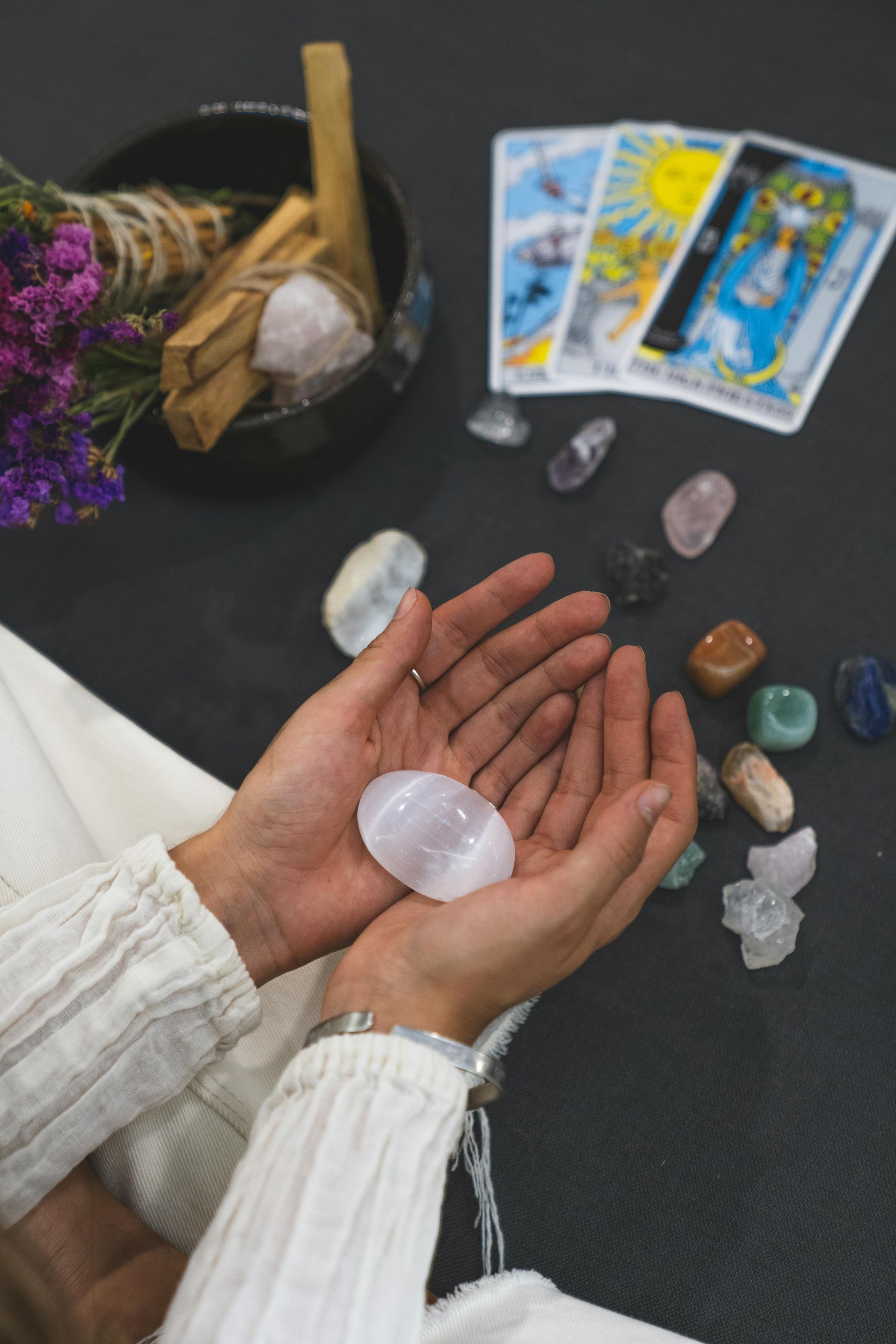 Hands holding a rose quartz, surrounded by crystals, tarot cards, and sage bundle on a dark surface.
