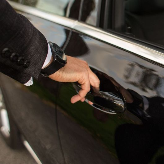 A man wearing a watch is opening the door of a black car