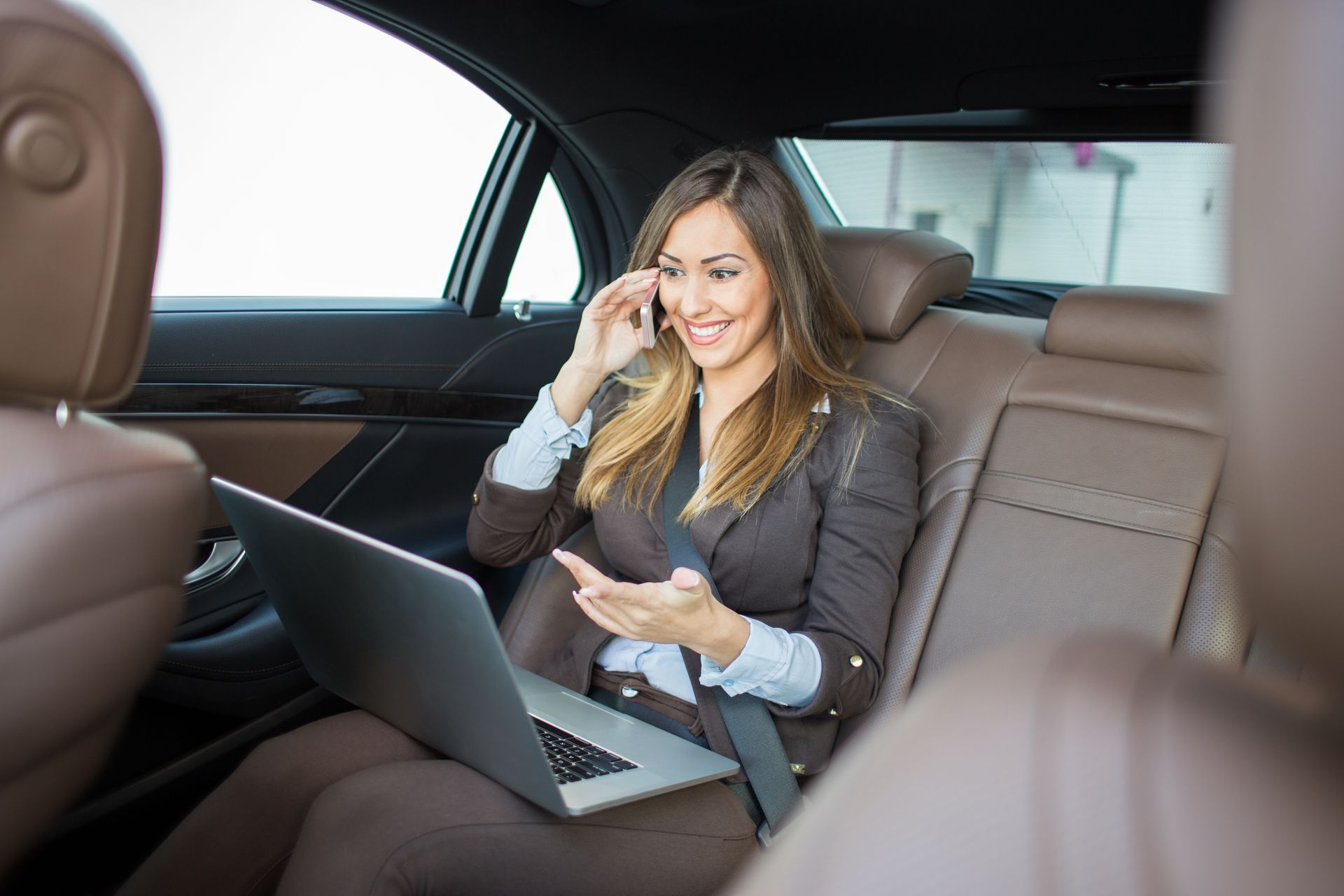 A woman is sitting in the back seat of a car using a laptop and talking on a cell phone.