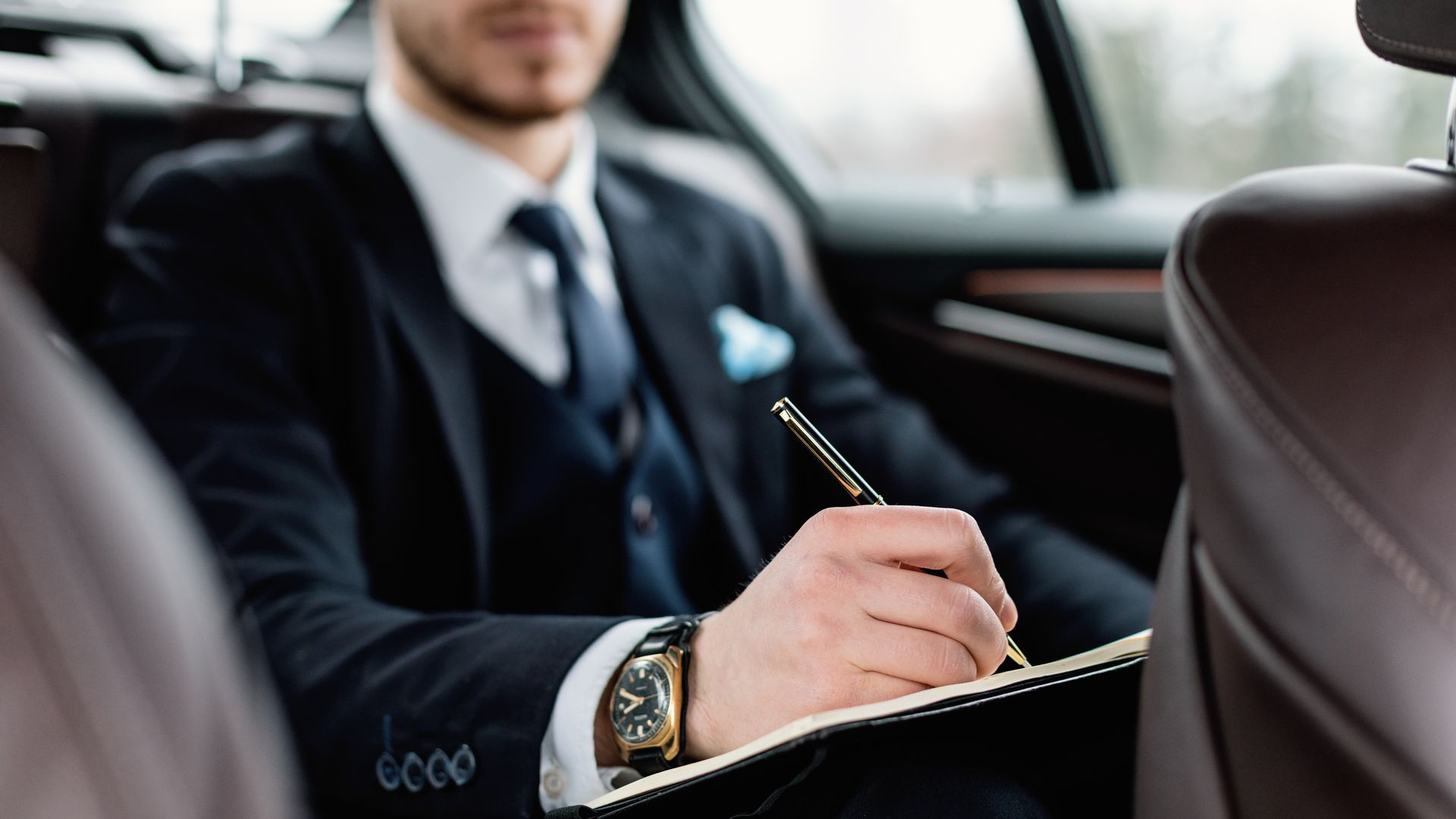a man in a suit and tie is sitting in the back seat of a car writing in a notebook .