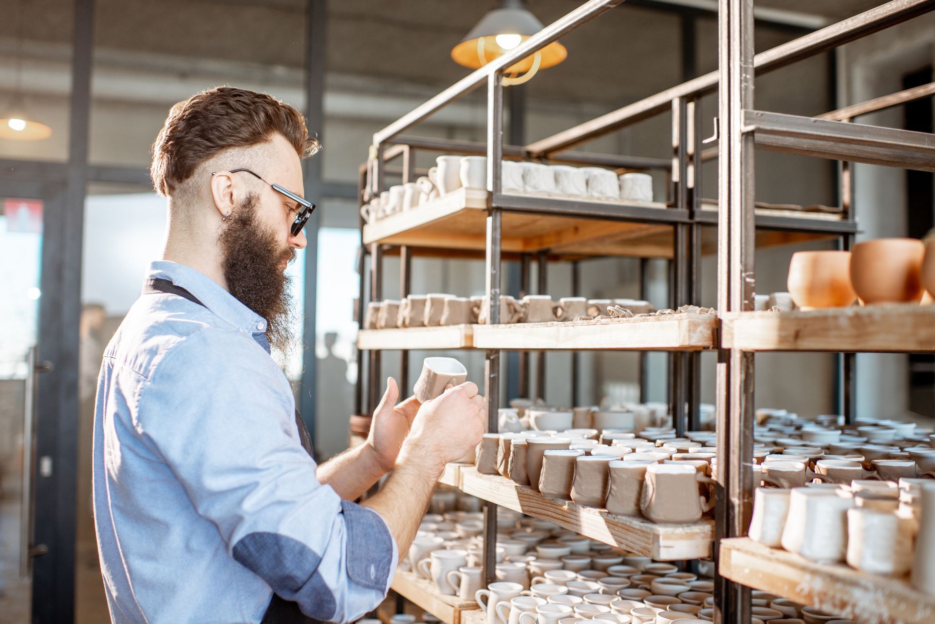 Man with beard inspects pottery on wooden shelves in a workshop.