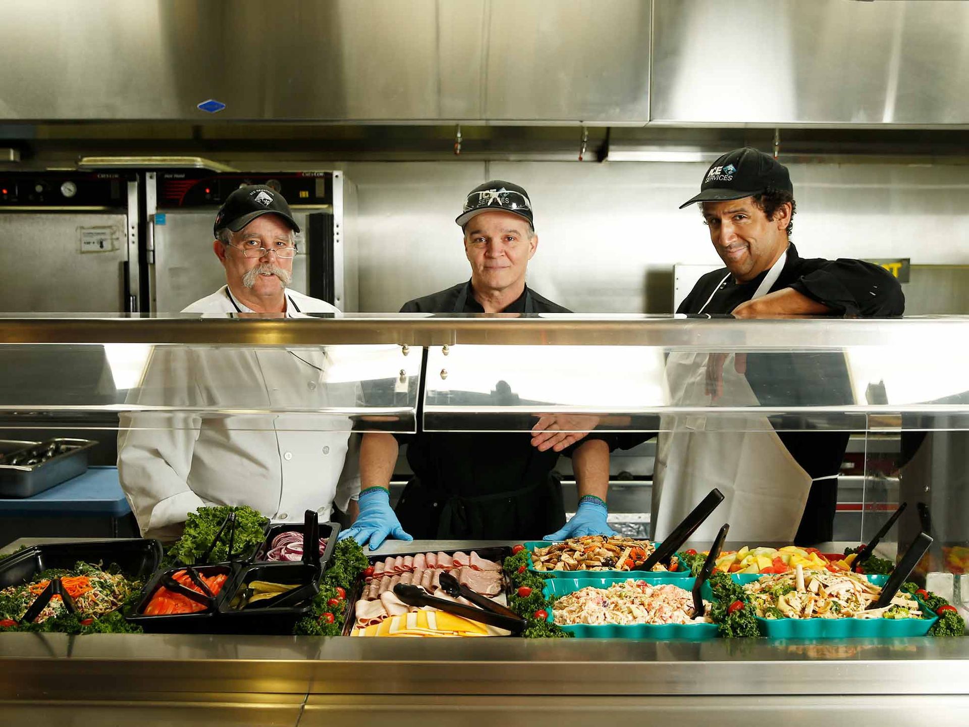 Three men are standing behind a buffet line in a kitchen