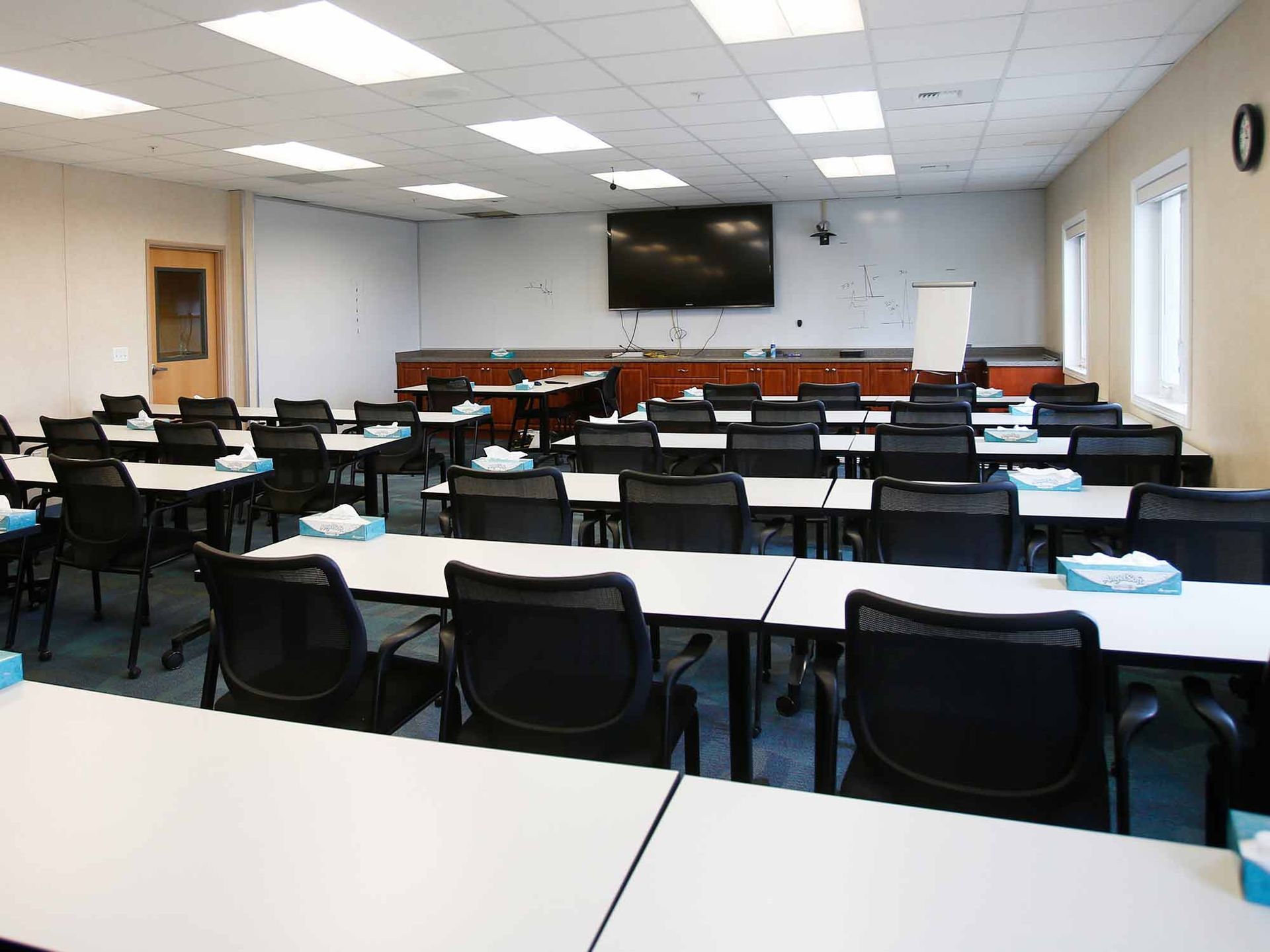A large classroom with tables and chairs and a flat screen tv on the wall.