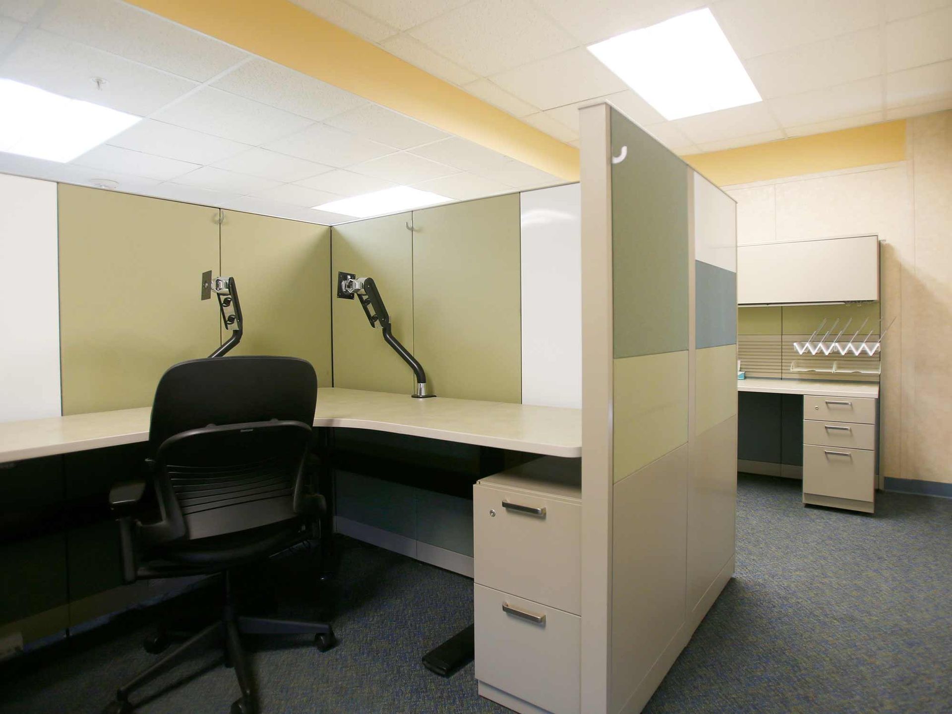 A cubicle in an office with two desks and a chair