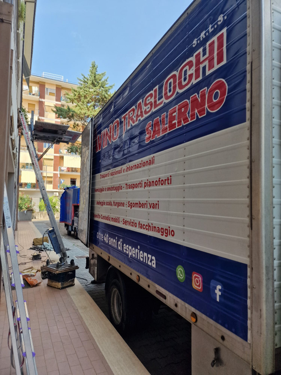 Un camion per traslochi blu e bianco è parcheggiato in una strada stretta, accanto a un marciapiede e a dei palazzi.