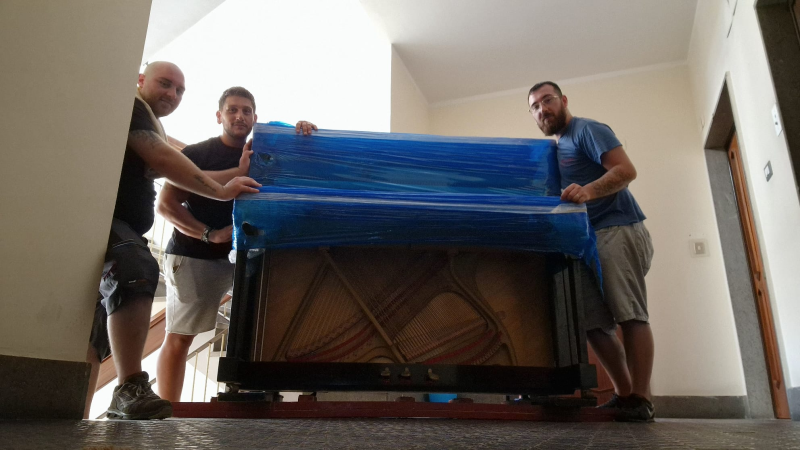 Tre uomini in un corridoio trasportano un pianoforte avvolto in un telo blu su un carrello mobile.