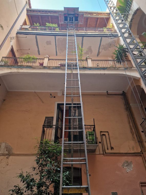 Una lunga scala a pioli si protende da un cortile verso un balcone di un edificio color pesca.