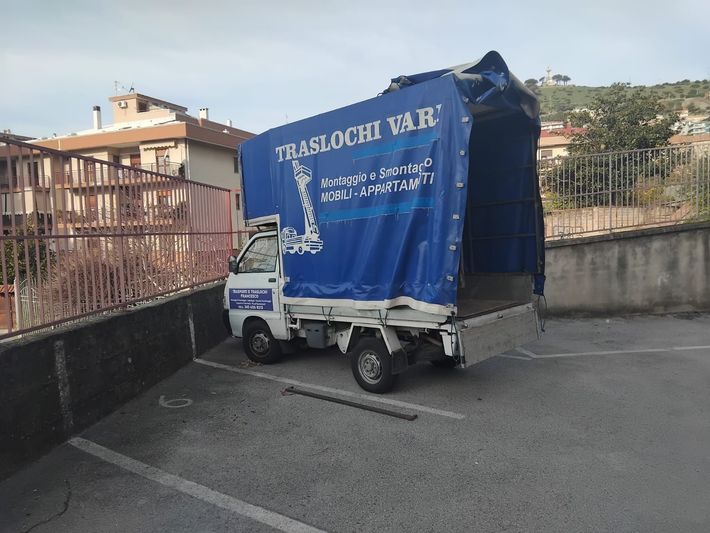 Un piccolo camioncino bianco con un cassone coperto di blu è parcheggiato in strada, accanto a un muro e ad alcune case.