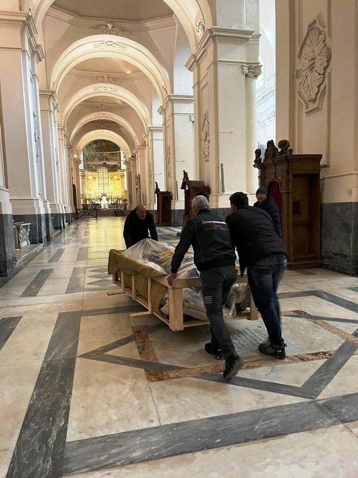 Persone che spostano una lunga panca di legno lungo la navata di una grande cattedrale.