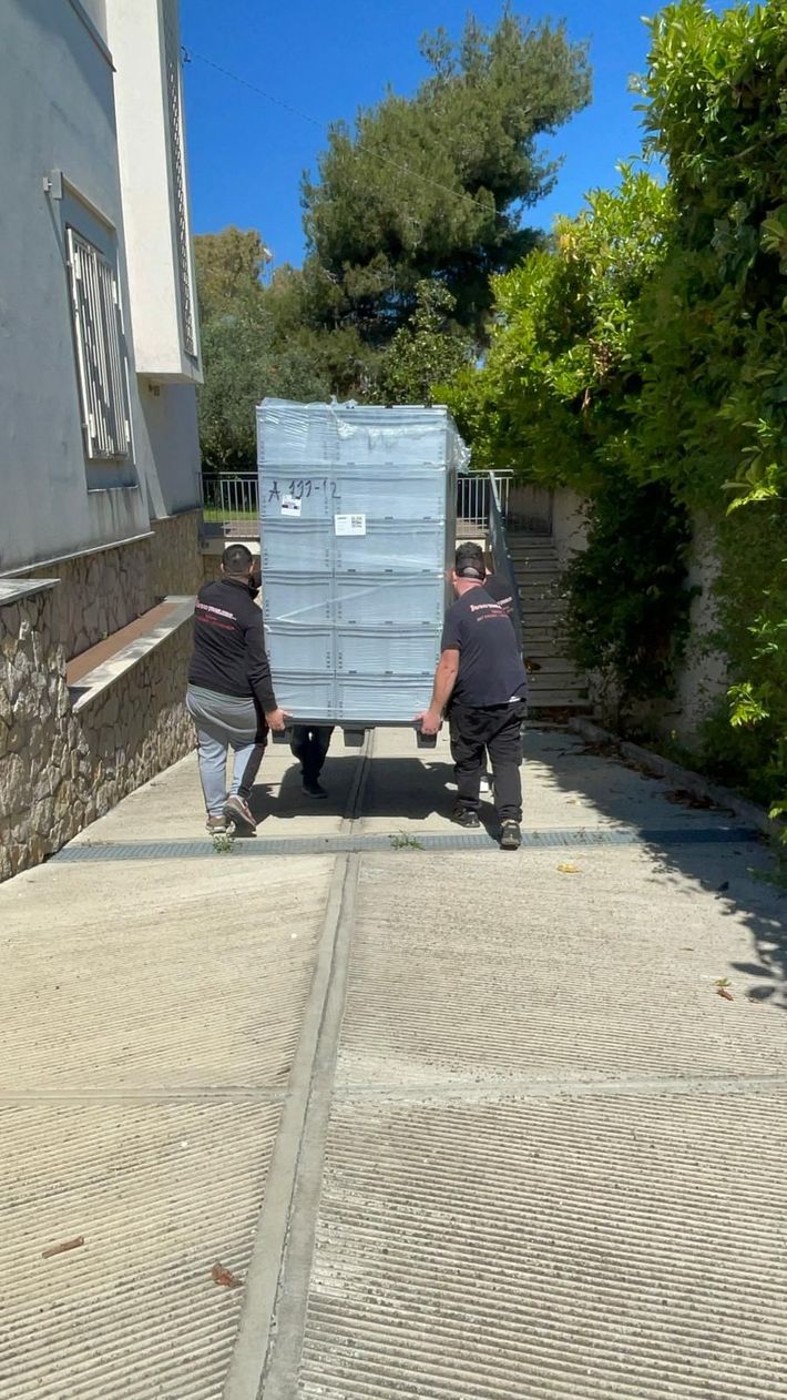 Due operai spingono un grande container metallico lungo un vialetto asfaltato e soleggiato tra gli edifici.