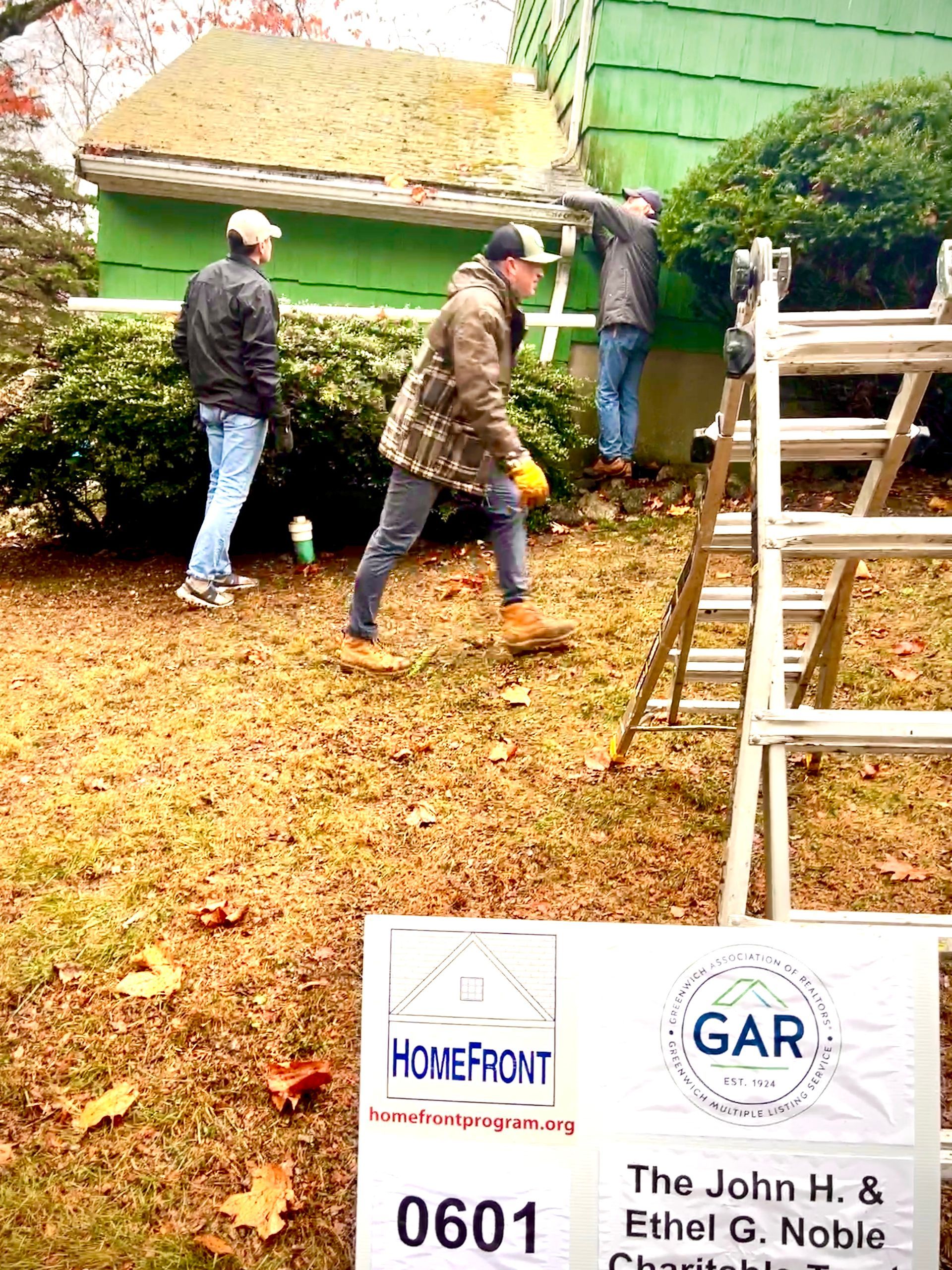 Three men working on a green house, one on a ladder. Sign for a charitable foundation in yard.