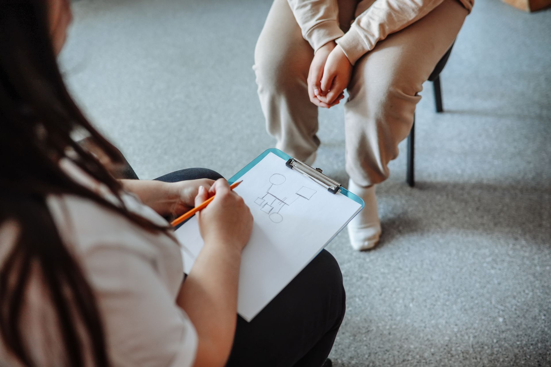 A woman is sitting in a chair talking to another woman while holding a clipboard.