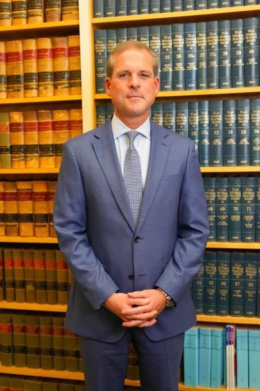 A professional in a blue suit and tie stands with hands clasped in front of a floor-to-ceiling wooden library bookshelf.